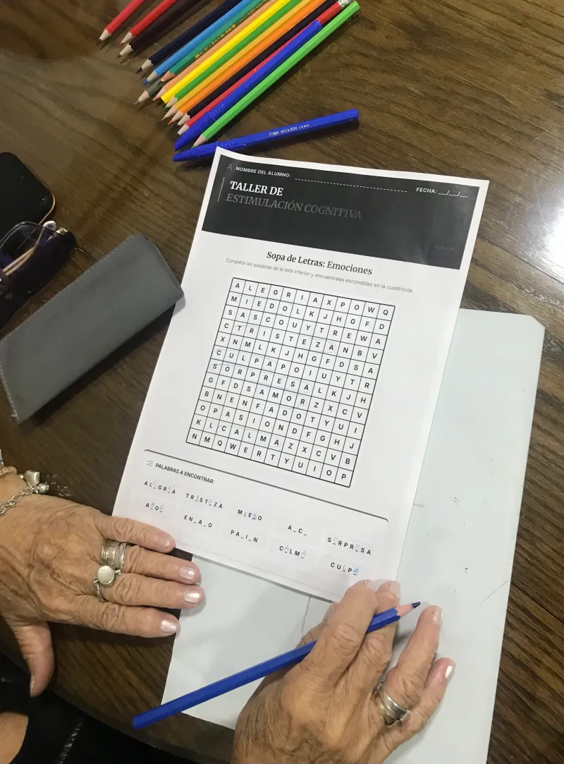 Manos de una alumna completando una sopa de letras sobre emociones, con lápices de colores sobre la mesa